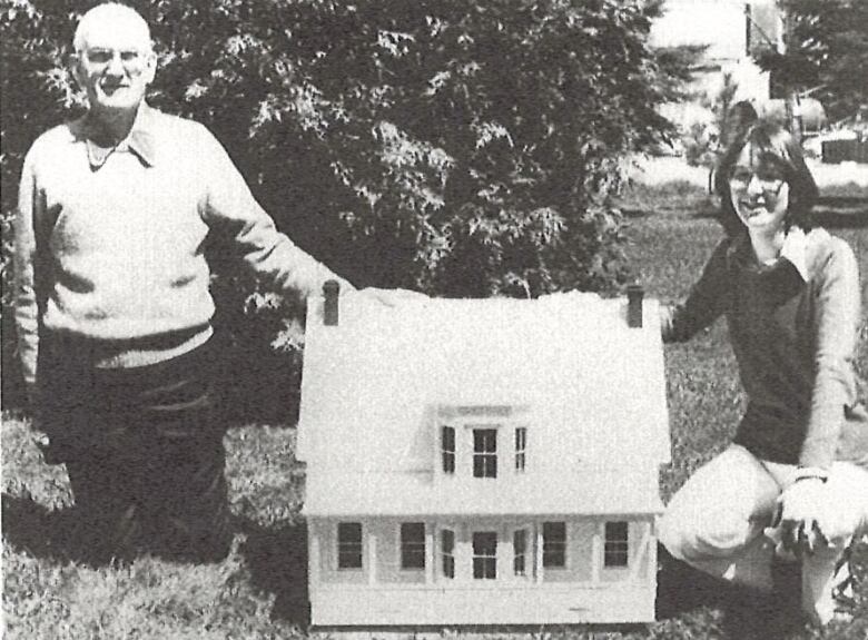 A black and white photo of an older man, left, and a younger woman, right, both kneeling next to a dollhouse.