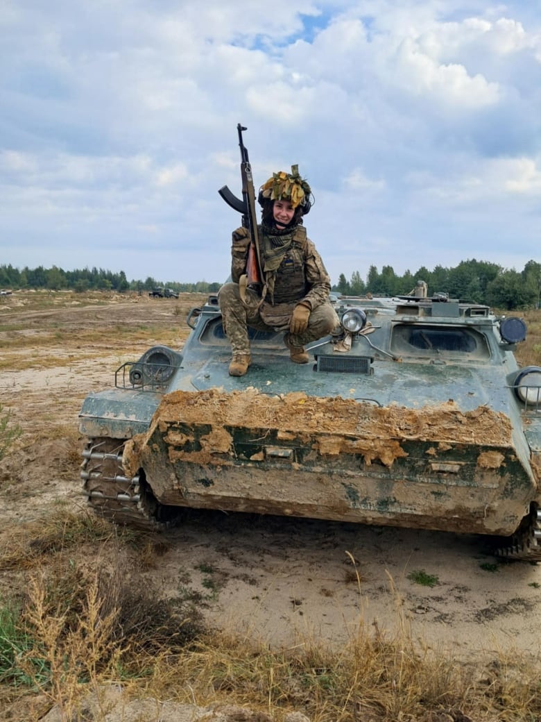 A woman in a military uniform holding a rifle, crouching over a tank.