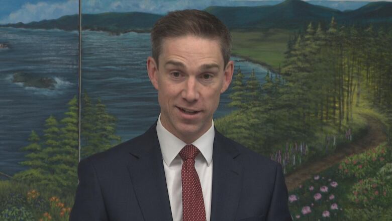 Man in blue suit and red spotted tie. Behind him is a mural of a painted shoreline of water and trees.