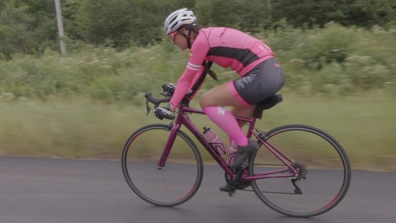 A woman in a pink cycling suit rides her bike on the side of an asphalt road. 