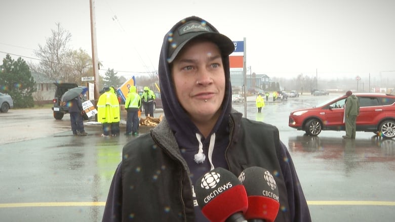 A woman wearing a dark hoodie and dark ballcap stands in front of microphones in the rain while people in neon yellow rain slickers stand in back.