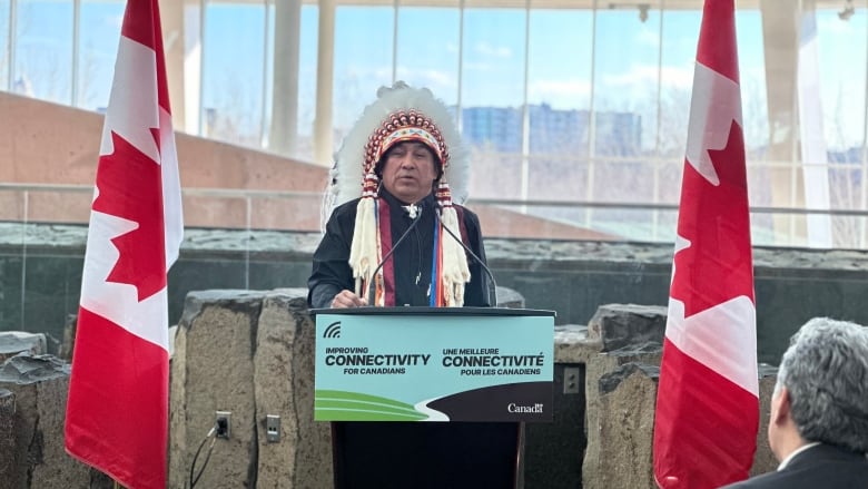 A man wearing a head dress stand being a podium and addresses a crowd.