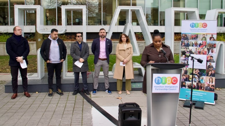 A line of people stand outdoors in front of a sign spelling out 