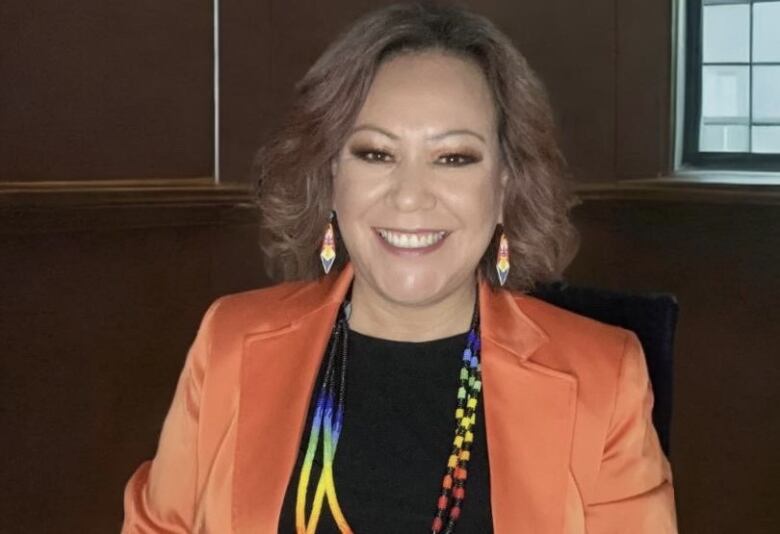 A woman sits for a photo in beaded earings and an orange blazer.