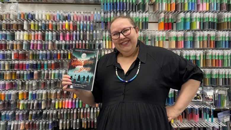 A woman is holding a book, standing in front of a wall of containers of colourful beads. 