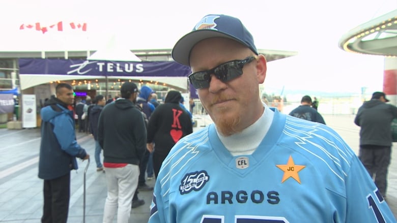 A fan in a blue jersey and sunglasses stands outside.