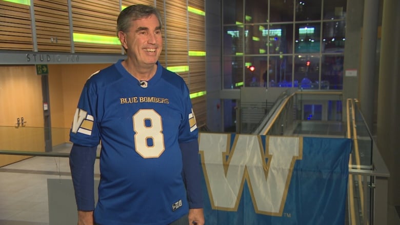 A man in a jersey stands next to a blue and white flag.