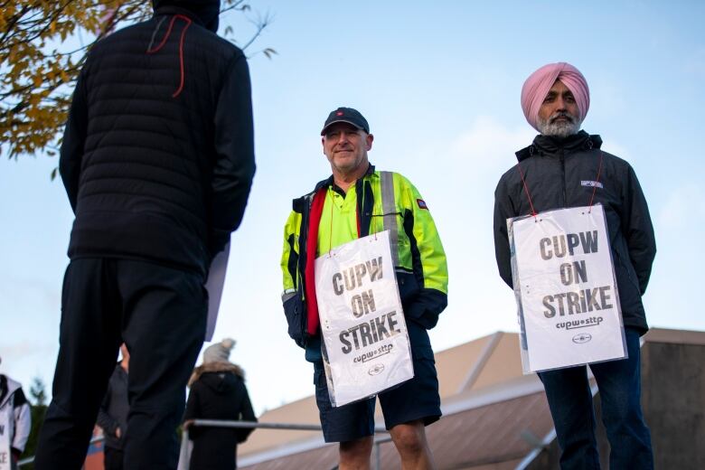 Two workers wear sandwich boards reading 'CUPW On Strike' outdoors.