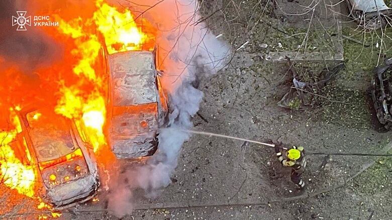 An overhead view shows a firefighter using a hose to douse burning cars.