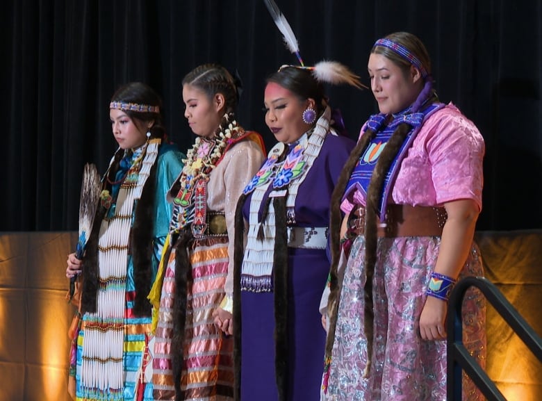 Four woman in traditional Indigenous clothing hold hands on a stage.