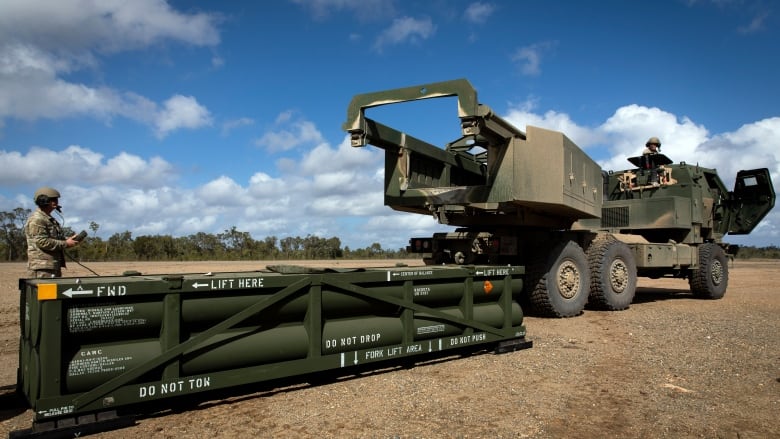 A military vehicle is shown on a dirt ground with a large weapon in a crate nearby. A soldier is also shown.