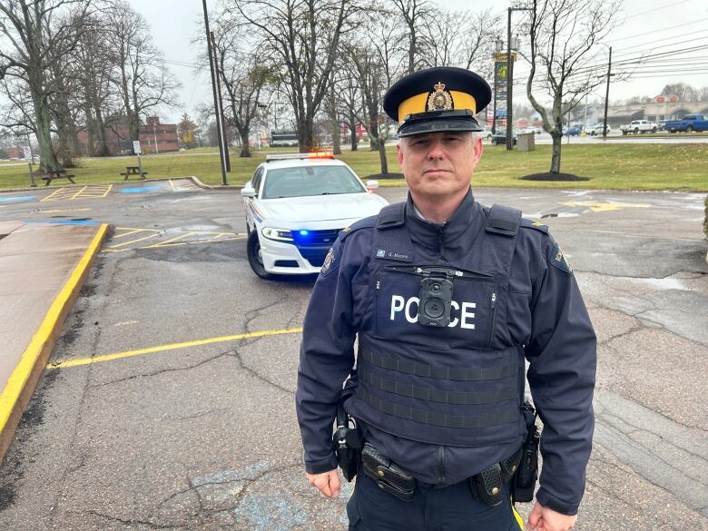 A male RCMP officer in uniform looking at the camera with a police cruiser with lights on behind him.