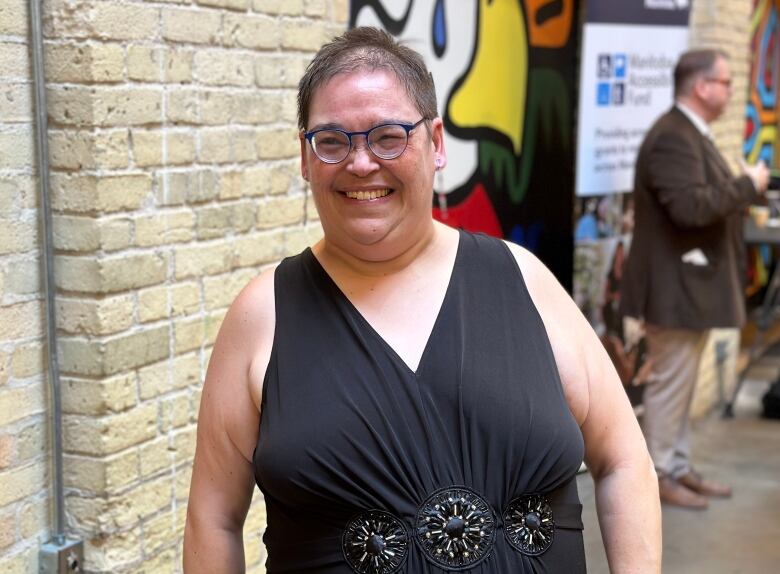 A woman wearing a black dress smiles in front of a brick wall. 