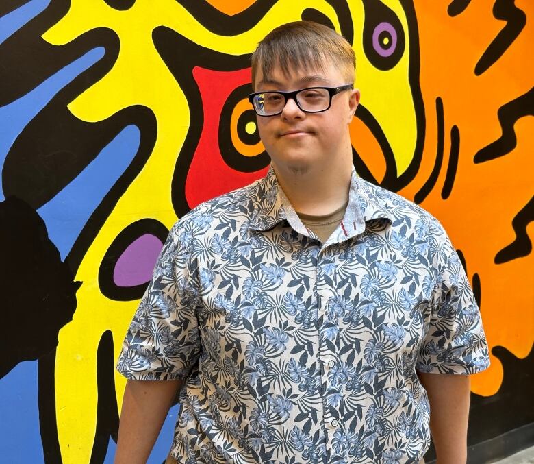 A man wearing a blue and white button up shirt standing in front of a colourful wall. 