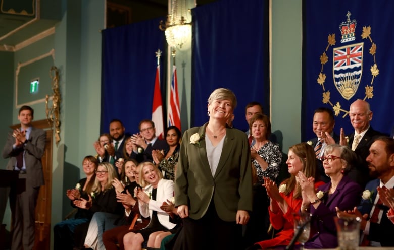 A woman stands up as a series of seated people applaud for her.