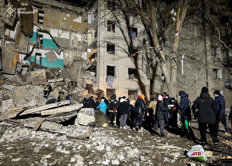 Dozens of people stand outside a multistorey residential building that has been partially destroyed, with concrete debris on the ground.