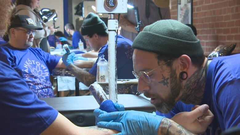 A man wearing a toque and glasses, with visible facial tattoos, is holding a tattoo gun to tattoo a man's arm.