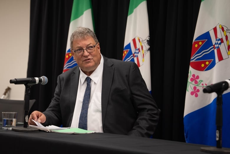 Man in suit at table with notes by microphone.