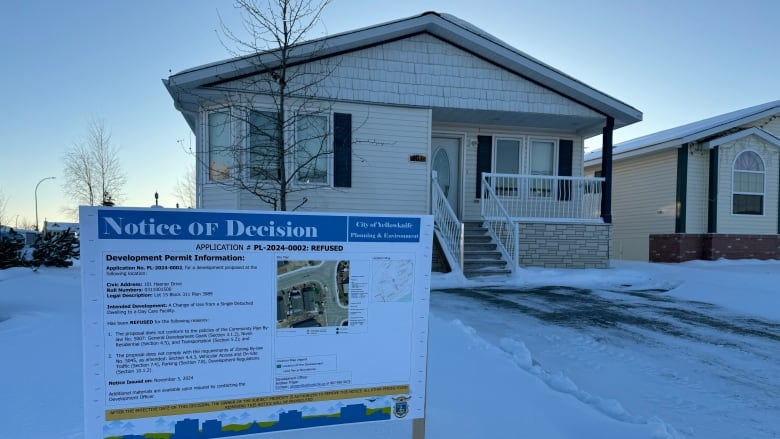 A board with writing on it outside a white home with a snow-covered lawn.