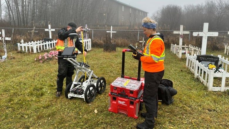 Two people in a cemetery with a red block-shaped machine. 