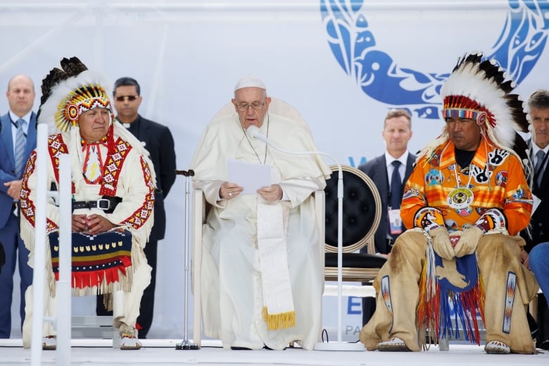 A man in white robes sits with two Indigenous men on either side of him.