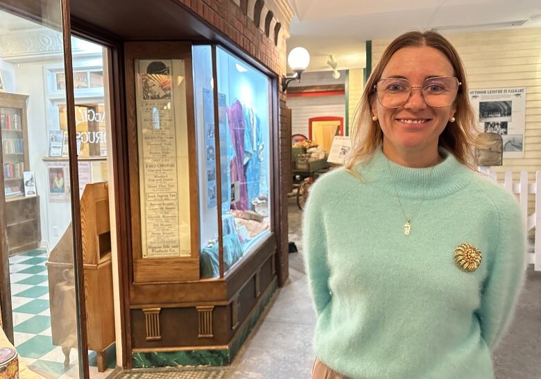 A woman smiles in a museum setting. She has light-coloured hair and is wearing a sweater and glasses.