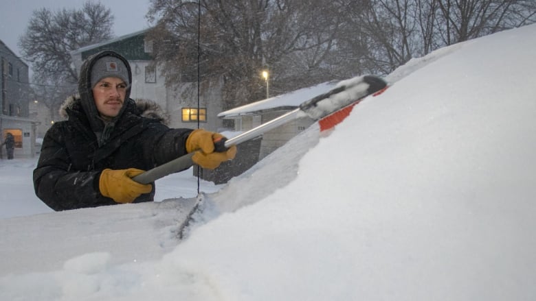 A man in a parka clears snow off the windshield of a car.