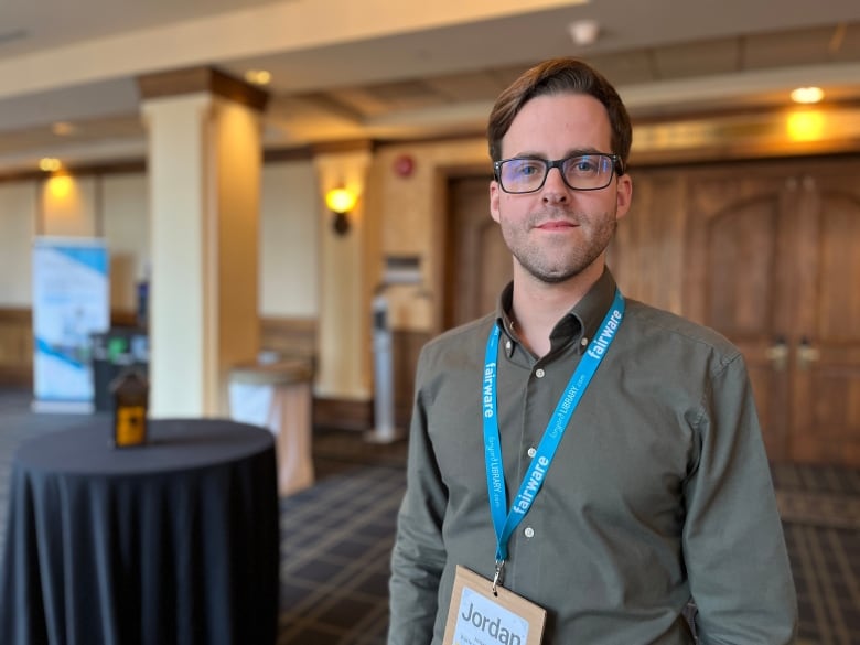 A man with brown hair and glasses, wearing an olive-grey shirt, is pictured inside a hotel conference centre.