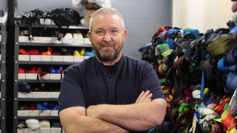 A man with short hair and a beard, wearing a navy blue t-shirt and fleece vest, poses with his arms crossed in front of a display of zippers.