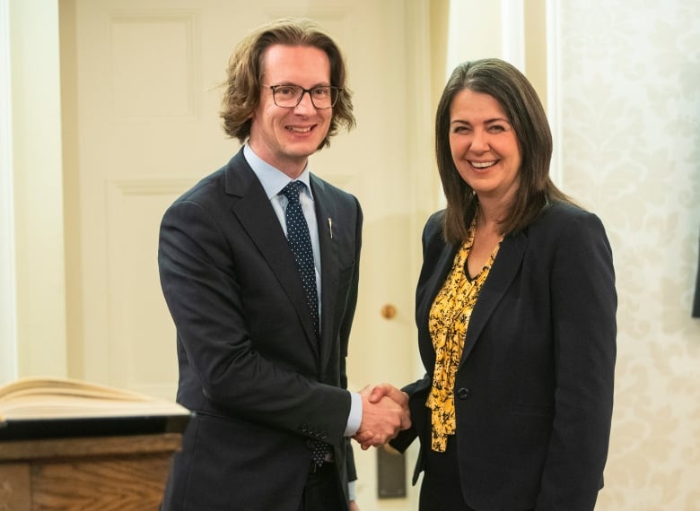 A man in a suit wearing glasses shakes hands with a woman wearing a suit.