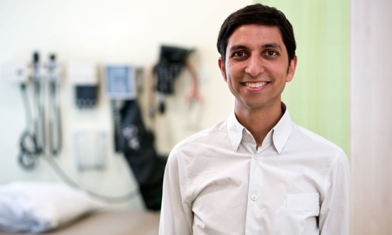 A clean shaven man with short, black hair wearing a white collared shirt stands in an examining room with a diagnostic set on the wall behind him.