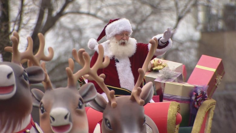 Santa Claus standing on a float with reindeer, waving to people below 