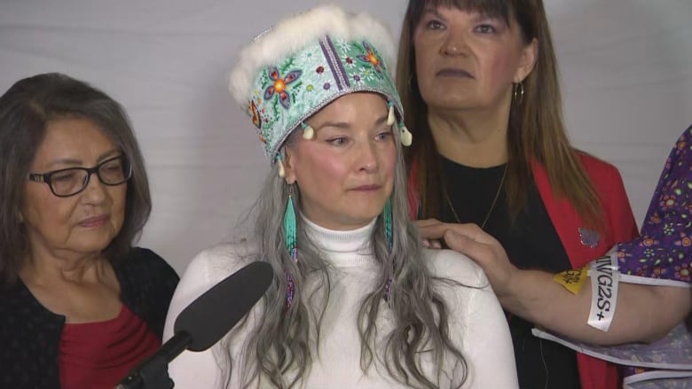 A woman wearing an Indigenous headdress speaks, while three other women stand behind her.