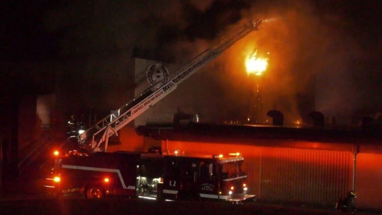 A fire truck with its ladder raised outside a burning building in the middle of the night.