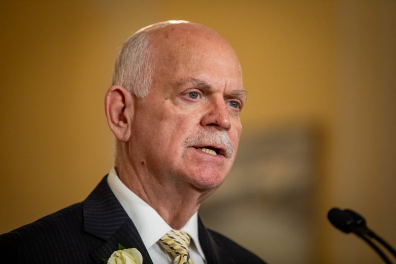 A white man with balding white hair speaks into a mic.