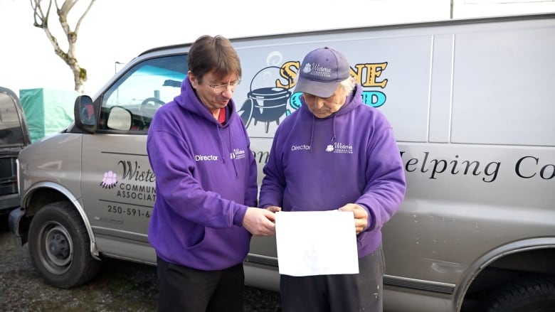 Two men wearing purple hoodies look at a receipt in front of a van.