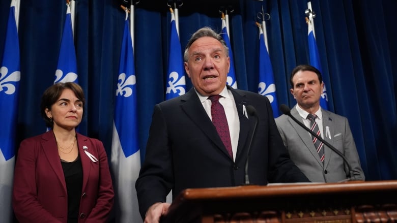 A middle aged white man speaks at a podium with Quebec flags behind him and a man and a woman on either side of him.