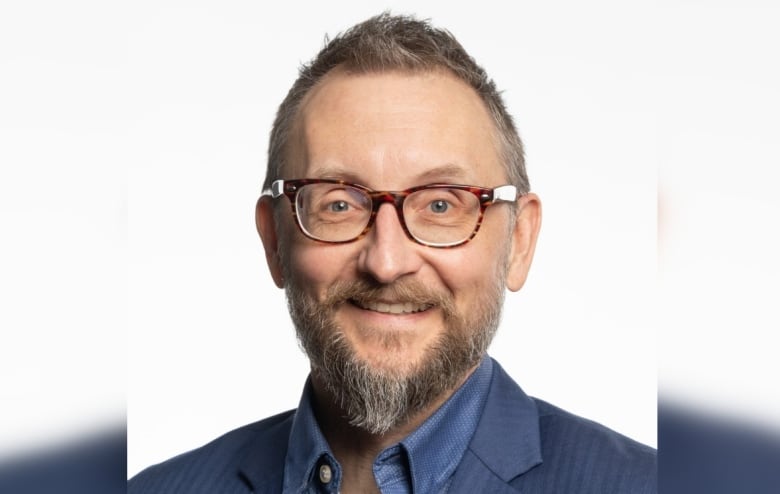 A man with short grey hair and a grey goatee is seen from the head and shoulders, smiling and wearing glasses.