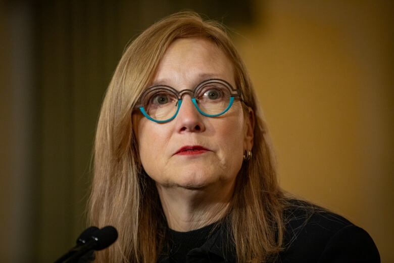 A middle-aged woman with long red hair with round blue-rimmed glasses looks straight ahead.