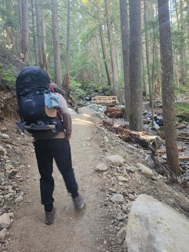 A man's backpack is pictured towards the camera as he stands on a wooded trail.