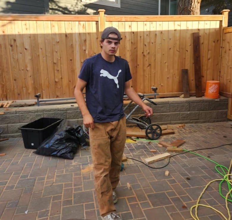 A man wearing a dark blue T-shirt and brown pants is pictured in front of a fence.